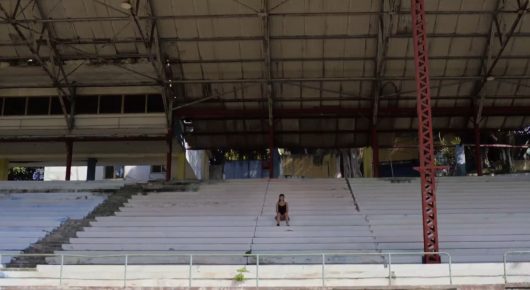 Still from Tricontinental: a nearly empty arena with a lone figure seated among rows of white stands under a high roof.