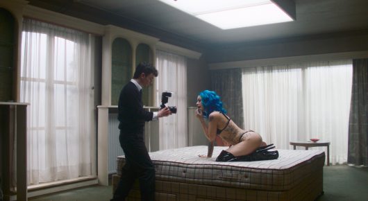 Still from Truly Naked: a camera operator films a person with blue hair kneeling on a mattress in a large room with tall windows.