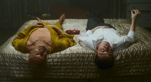 Still from Truly Naked: two people lie on their backs on a bed with a patterned cover, seen from above, one in a yellow top.