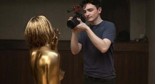 Still from Truly Naked: a man holding a video camera stands beside a person whose body is painted gold.