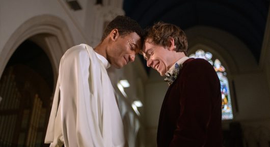 Still from Cambridge Connections: two men in period costume, one in a white robe and one in a dark velvet jacket, lean their foreheads together in a church.