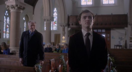 Still from Cambridge Connections: two men in dark suits stand apart in the aisle of a church, looking towards each other.