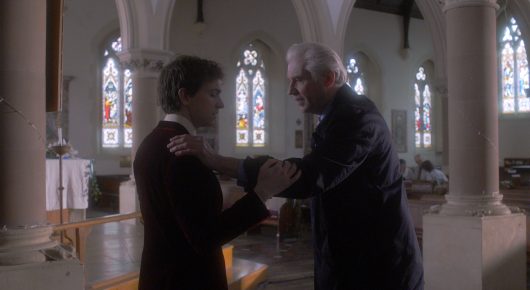 Still from Cambridge Connections: two men in dark suits shake hands in a church, one younger and one older.
