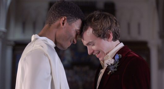 Still from Cambridge Connections: two men in period costume, one in white and one in dark velvet, press their foreheads together in a church.