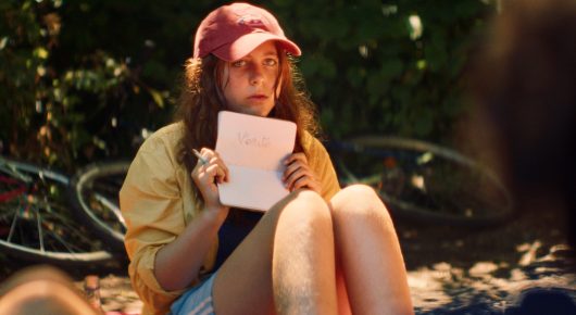 Still from The Harvest: a girl in a red cap and yellow jacket sits on the grass holding a sheet of paper, with a bicycle behind her.