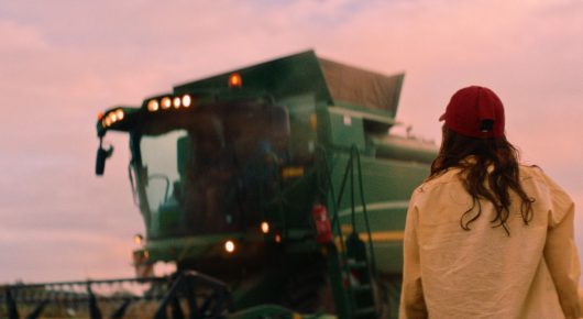 Still from The Harvest: a girl in a red cap and yellow jacket, seen from behind, stands facing a green combine harvester at sunset.