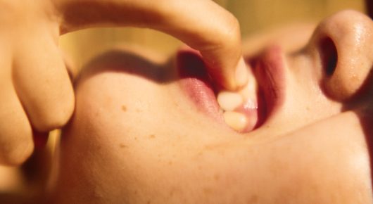 Still from The Harvest: an extreme close-up of a mouth with a finger at the lips.