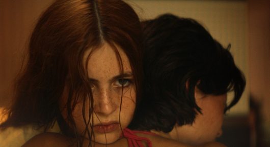 Still from The Harvest: a girl with red hair and wet skin looks at the camera, close to another person whose head is turned away.