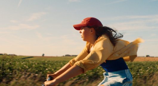 Still from The Harvest: a girl in a red cap and yellow top rides a bicycle fast across a field, her hair blowing back.