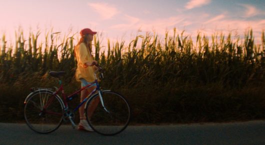 Still from The Harvest: a girl in a red cap rides a bicycle along a road beside a field of tall crops at sunset.