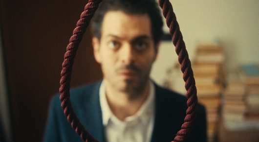 Still from The Last Ingredient: a man in a navy jacket and white shirt looks at the camera with a looped rope hanging in front of him.