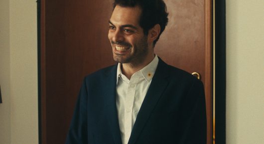 Still from The Last Ingredient: a smiling man in a dark jacket and white shirt stands in a doorway.