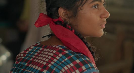 Still from The One Who Plays: a close-up of a girl with a plait, a red scarf and a checked shirt, looking off to one side.