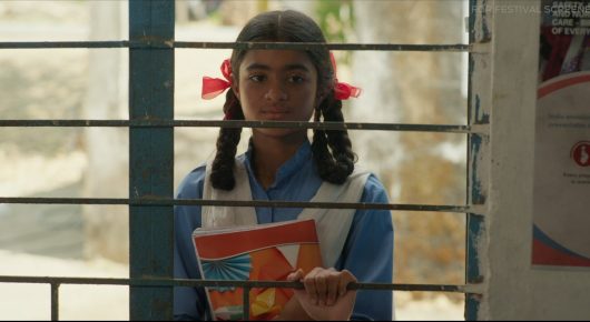 Still from The One Who Plays: a girl in a blue school uniform with red hair ribbons holds a book, standing behind a barred window.