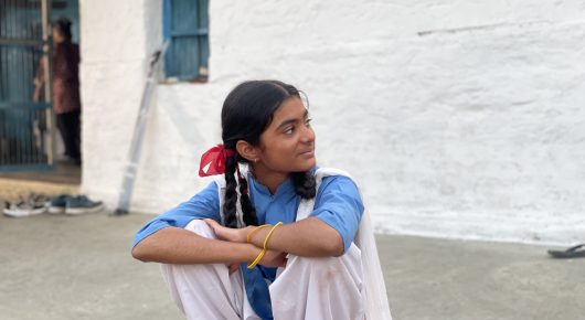 Still from The One Who Plays: a girl in a blue top with plaits and red hair ties sits with her arms around her knees outside a white building.
