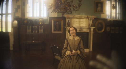 Still from The Painting and The Statue: a woman in a Victorian-style dress stands very still in a wood-panelled room with sunlight through tall windows.