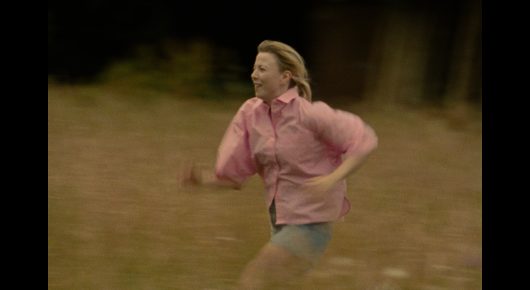 Still from The Pigeon: a woman in a pink jacket runs across a grassy field.