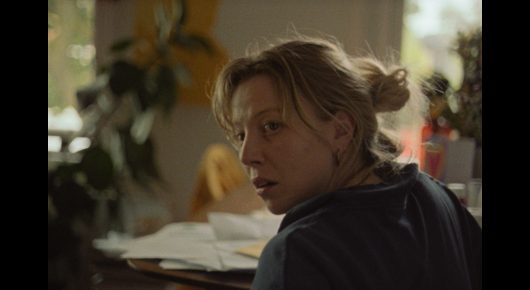 Still from The Pigeon: a woman with her hair in a bun looks back over her shoulder at a cluttered desk by a window.
