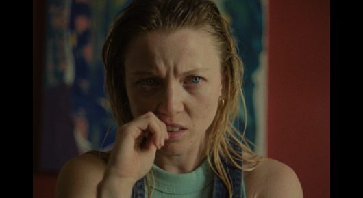 Still from The Pigeon: a woman with damp blonde hair covers her mouth with her hand and frowns, in front of a red wall.