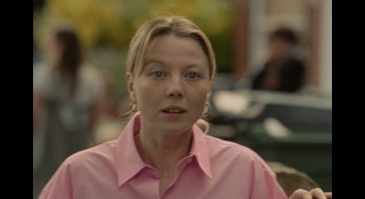 Still from The Pigeon: a woman in a pink collared shirt looks slightly to one side, with a blurred busy room behind her.