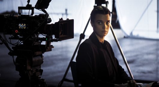 Still from The Secret Assistants: a woman sits beside a film camera on a tripod, with a hazy bright background.