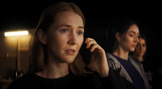 Still from The Secret Assistants: a woman with long auburn hair holds a phone to her ear, with another woman behind her.