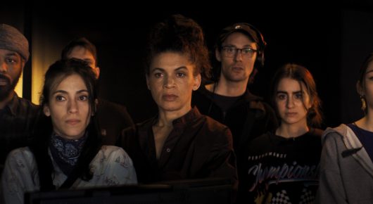 Still from The Secret Assistants: a group of people, one wearing a headset, look towards something off-screen in the dark.