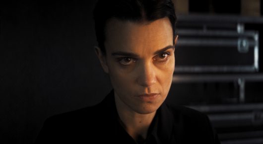Still from The Secret Assistants: a close-up of a woman with short dark hair staring at the camera, with film equipment cases behind her.