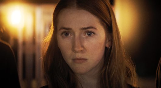 Still from The Secret Assistants: a woman with long auburn hair looks at the camera in warm light, with a blurred street light behind her.