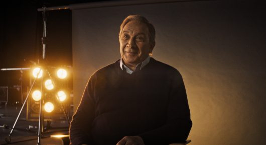 Still from The Secret Assistants: a smiling older man in a dark jumper stands in front of studio lamps.