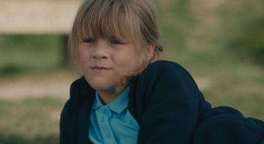 Still from To Catch a Siren: a child with blonde hair in a dark cardigan lies on the grass, looking at the camera.