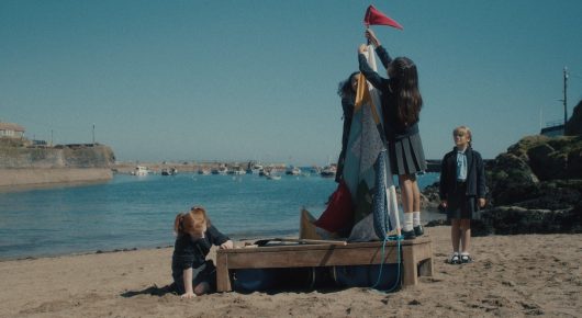 Still from To Catch a Siren: a tall figure in a long dark coat and others stand on a beach next to a wooden boat with a red flag, with a harbour in the distance.