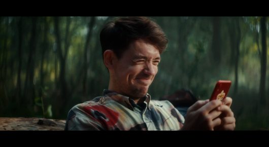 Still from Todd and The Toad: a smiling man in a patterned shirt looks at a phone in his hand, in a wood.