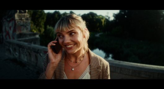 Still from Todd and The Toad: a woman with short blonde hair talks on a phone beside a stone balustrade, with trees behind her.