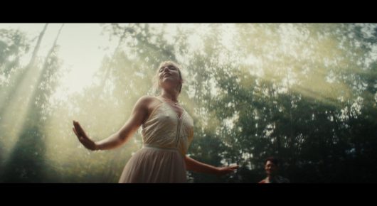 Still from Todd and The Toad: a woman in a pale dress stands with her arms outstretched in a sunlit forest.