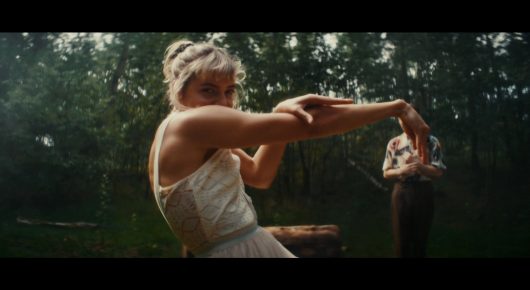 Still from Todd and The Toad: a woman in a pale dress with her hair in a bun stretches out an arm in a forest clearing, with a man standing behind her.