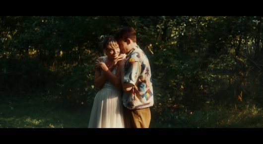 Still from Todd and The Toad: a man in a patterned shirt and a woman in a pale dress embrace in a green clearing.