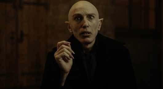 Still from Uncle Artoo: a pale, bald man in a long black coat raises one hand near his face and looks wide-eyed at the camera in a dim room.