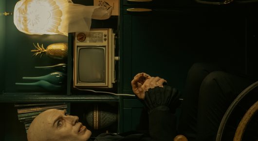 Still from Uncle Artoo: a bald man lies on a sofa in a cluttered green room with a vintage television and a glowing lamp.