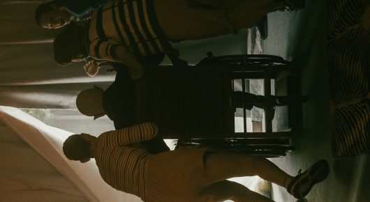 Still from Uncle Artoo: several people sit and stand around a chair in a dim room.
