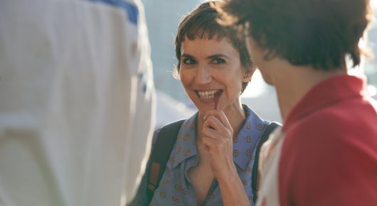 Still from Viva: a woman with short dark hair smiles with her hand at her chin, beside a person in a red jacket.
