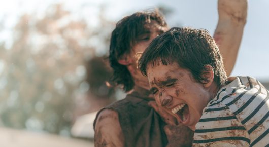 Still from Viva: two young men, one in a striped top, laugh together outdoors with dirt on their faces.