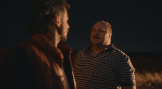 Still from Vultures: two men talk outdoors at night, one in a brown jacket and one in a striped top.