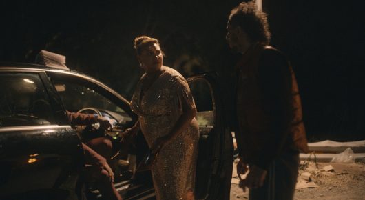 Still from Vultures: people stand talking beside a parked car at night.