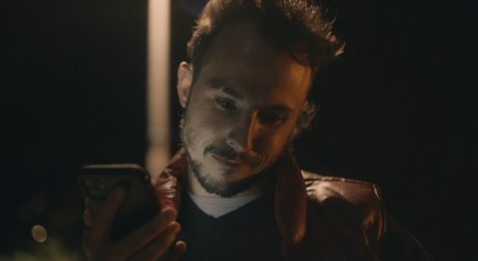 Still from Vultures: a man in a brown jacket looks down at a phone at night.