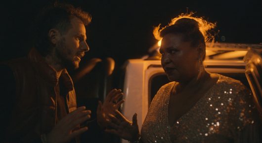 Still from Vultures: a man and a woman in a sequinned top talk beside a vehicle at night.