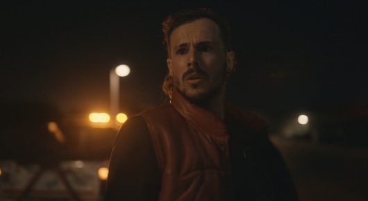 Still from Vultures: a man with light stubble in a red jacket looks off to one side at night, with street lights behind him.