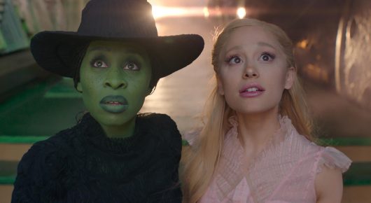 Still from Wicked: a green-skinned woman in a black pointed hat and a blonde woman in a pale pink dress sit side by side, looking up.