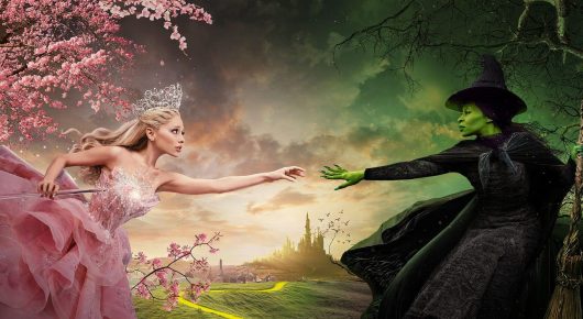 Still from Wicked: a woman in a pink gown and a witch in black reach towards each other over a fantasy landscape with pink blossom and green fields.