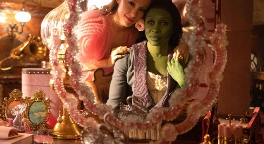 Still from Wicked: a green-skinned woman and another woman look into an ornate pink mirror in a decorated room.
